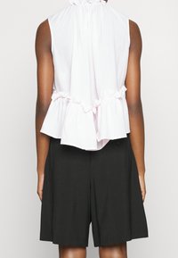 Blusa sin mangas de color rosa claro con un dobladillo con volantes y caída en la parte posterior, tejido texturizado; combinada con pantalones cortos negros a medida.