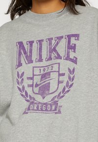 Gray sweatshirt featuring a large purple "NIKE" logo, 1972 text, and an emblem with laurel leaves and "OREGON" below. Soft cotton material.