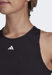 Czarny sportowy top na ramiączkach z prążkowanym dekoltem i białym logo Adidasa na górze po lewej stronie. Gładka tekstura materiału, bez rękawów.