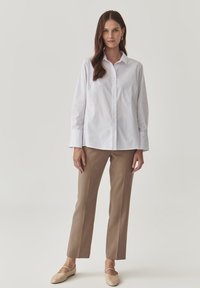 Camicia bianca con bottoni e maniche lunghe, abbinata a pantaloni beige a gamba dritta e scarpe slip-on beige chiaro.