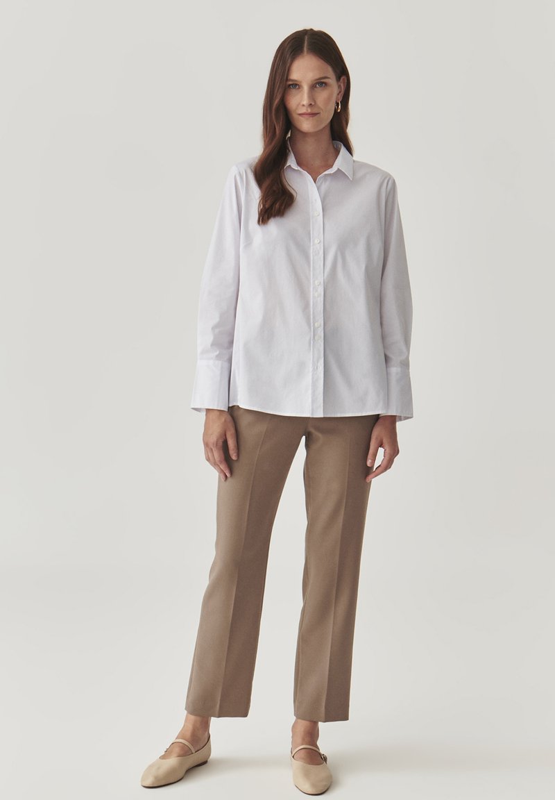Camicia bianca con bottoni e maniche lunghe, abbinata a pantaloni beige a gamba dritta e scarpe slip-on beige chiaro.