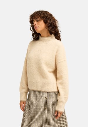 Femme aux cheveux bouclés portant un pull à col roulé crème et une jupe à carreaux noir et blanc boutonnée, debout devant un fond uni.