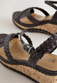 Įsispiriami sandalai su tamsiai rudos spalvos pintais odos dirželiais ir kamštiniu padu, viename dirželyje yra reguliuojama metalinė sagtis.