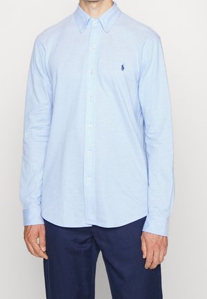 Mężczyzna w jasnoniebieskiej koszuli z długim rękawem zapinanej na guziki z kołnierzykiem oraz w granatowych spodniach, stojący na białym tle.