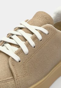 Beige suedinesko med hvite snøring, sydd detaljer, polstret krage og en lys brun gummisåle. Teksturert overflatestruktur.