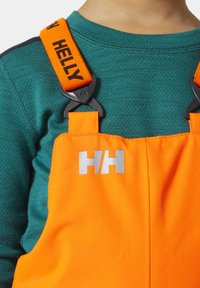 Oranžové voděodolné overal s černými detaily, s nastavitelnými popruhy. Na přední straně je vytištěno logo "HELly" v černé a stříbrné barvě.