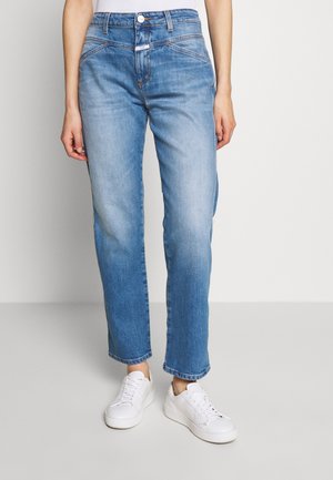 Persoon die lichtblauwe, rechte jeans met hoge taille en witte sneakers draagt, staand tegen een effen lichte achtergrond.