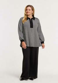 Schwarz-weiße Hahnentrittmuster-Tunika mit schwarzem Kragen und Ärmeln, kombiniert mit weiten schwarzen Hosen. Sie hat einen halben Reißverschluss am Hals und eine lockere Passform.