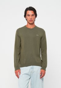Olijf groene gebreide trui met lange mouwen, ronde halslijn en geribbelde zoom. Voorzien van een klein logo op de linkerborst. Gecombineerd met lichtblauwe jeans.