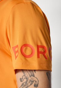 Oranžinė trumpa rankovė marškiniai su raudonais žodžiais ant rankovės ir matomu roplio tatuiruotė ant apatinės rankos.