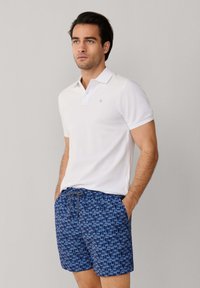 Homme portant un polo blanc à manches courtes et un short bleu à motif avec cordon de serrage, debout les mains dans les poches devant un fond uni.