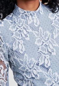 Vêtement en dentelle bleu clair avec des motifs floraux et un col haut, doté d'une base texturée et de manches transparentes. Les accents comprennent des détails complexes.
