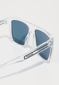 Lunettes de soleil polarisées avec monture transparente, verres teintés foncés, design angulaire et accent logo noir sur la branche.