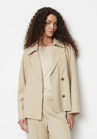 Beige kaheklöideline jakk laia kraega, rinnatasku ja vaba lõikega, valmistatud pehmest kangast. K-and ja valge ülemise osa kohal.