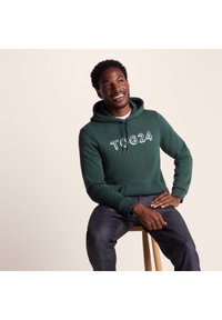 Green cotton hoodie with drawstring, featuring white "TOG24" logo, worn with dark denim jeans, seated on a wooden stool.