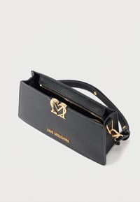 Μαύρη υφασμάτινη δερμάτινη τσάντα Love Moschino με χρυσό λογότυπο, καρδιόσχημο κλείσιμο, τσέπη με φερμουάρ και ρυθμιζόμενο λουρί ώμου σε απλό φόντο.