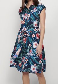 Kurzärmliges, knöpfiges Kleid aus marinefarbener Stoff mit floralem Muster, das rote und weiße Blumen sowie grüne Blätter zeigt und in der Taille gebunden ist.