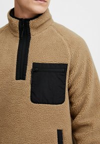 Suéter de polar color beige con un bolsillo en el pecho con cremallera negra y un cuello negro en contraste, con un acabado texturizado y cierre de medio zip.