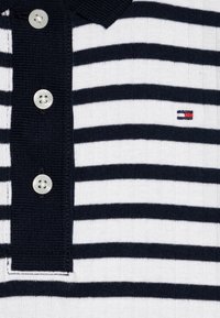 Pruhované polo tričko v navy a bílé barvě, s žebrovaným límečkem, knoflíkovým zapínáním a malým vyšitým logem s červenými akcenty.