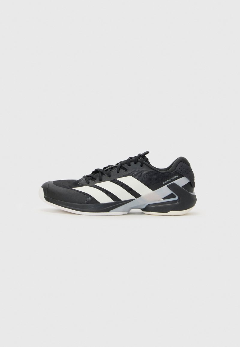 Chaussure de sport noire avec des rayures blanches, tige en mesh, bout arrondi et semelle rembourrée. Comprend un talon texturé et un système de laçage sécurisé.