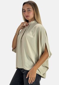 Beige, strukturiertes Fledermausshirt mit hohem Kragen, Seitenschlitzen und weiter Passform. Kombiniert mit dunkler Jeans, mit minimalistischem Design.