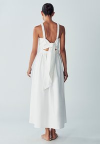 Weißes, ärmelloses Kleid mit einem Rückenschlitz und einer Schleifendetail an der Taille. Hergestellt aus leichtem Stoff, verfügt es über einen fließenden, knöchellangen Rock.
