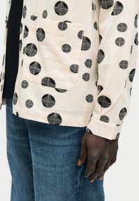 Μπεζ πουκάμισο με μαύρα κυκλικά σχέδια, τσέπη στο στήθος, φορεμένο με μπλε τζιν και ένα σκουρόχρωμο χέρι να ακουμπάει στο πλάι.