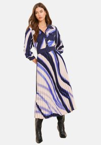 Plissiertes Midikleid in Blau und Creme mit gestreiftem Muster, Kragen, knöpfbaren Brusttaschen und tailliertem Gürtel. Lange Ärmel und fließender Stoff.