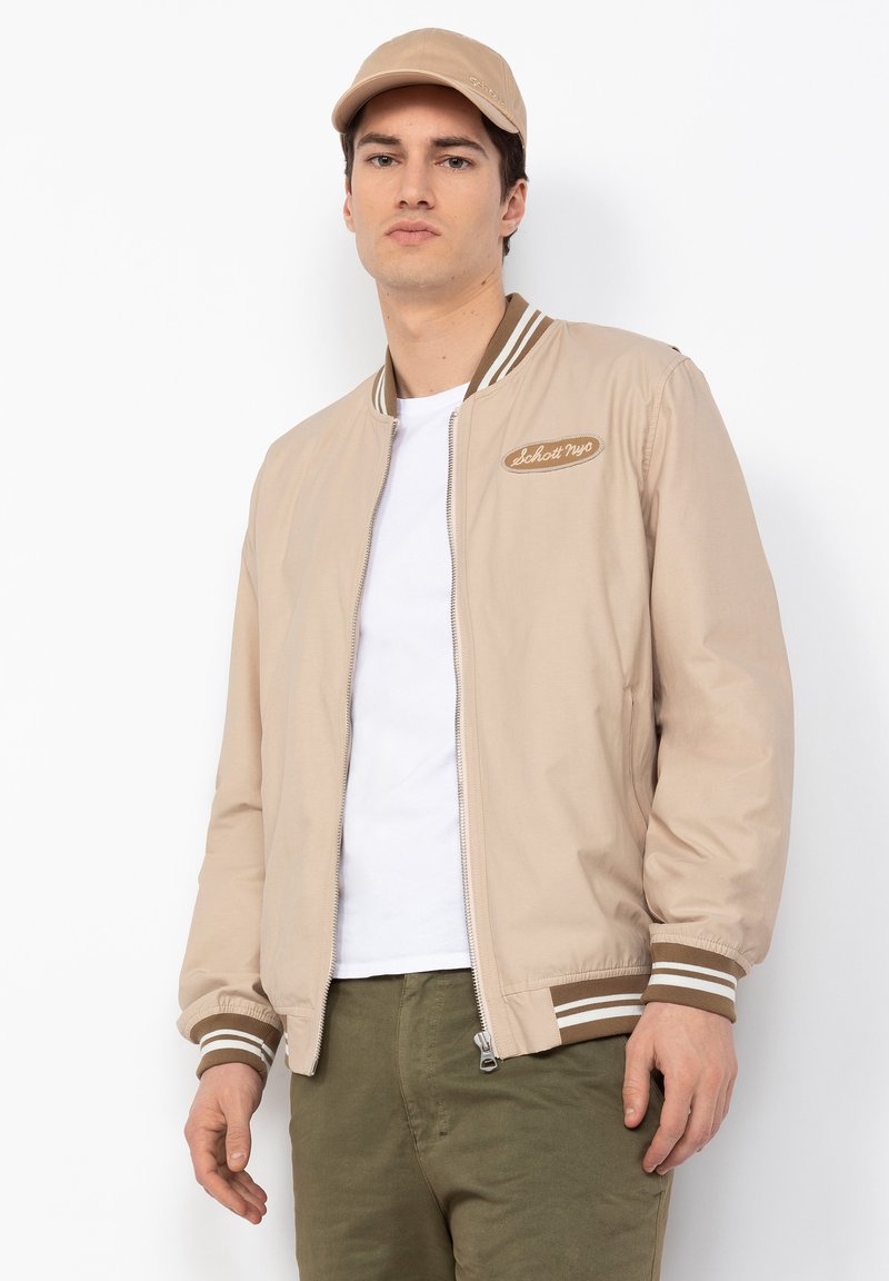 Beige Bomberjacke mit weißen und braunen gestreiften Rippbündchen und Kragen, mit einem Logo-Patch. Kombiniert mit einem weißen T-Shirt und Olivenhosen.