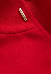Gros plan d'un tissu en laine rouge avec une couture diagonale et un petit ornement rectangulaire en or brillant.