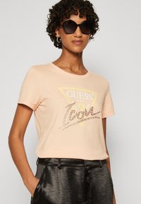  Lichtroze katoenen t-shirt met een ronde halslijn en korte mouwen, met een glanzend "GUESS" logo en een gele driehoekige accent.