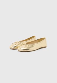 Ζευγάρι γυαλιστερές χρυσές μπαλαρίνες με μικρά διακοσμητικά φιόγκους στην περιοχή των δαχτύλων, απεικονισμένες σε απλό ανοιχτό γκρι φόντο.