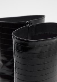 Czarne botki za kostkę z błyszczącą, skórzaną teksturą w wzór krokodyla. Posiadają zamek błyskawiczny i gładkie krawędzie. Model z otwartym wierzchem.