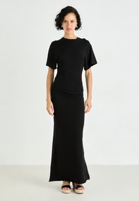Robe maxi noire avec des manches courtes évasées, un corsage ajusté et une texture lisse. Portée avec des sandales à plateforme noires présentant des détails tissés.