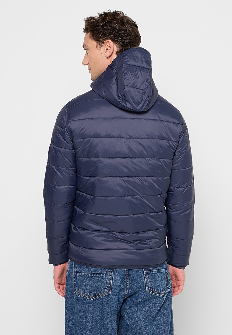 Granatowa kurtka puchowa z kapturem, poziomo pikowana i o błyszczącym wykończeniu. Noszona z dżinsami, prezentująca swobodny styl.