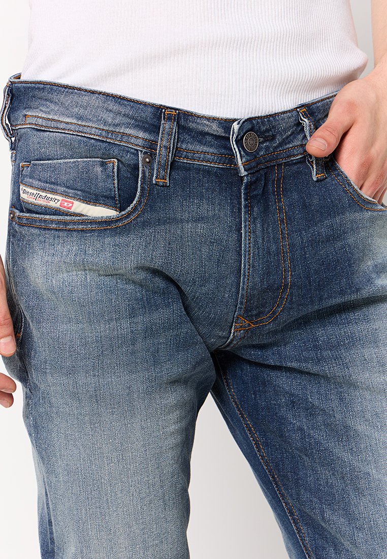 Jeans in denim blu scuro con un fronte sbiadito, cuciture arancioni e una piccola tasca laterale con etichetta. Chiusura con bottone e zip.