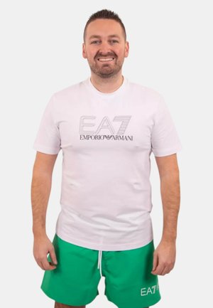 Uomo sorridente che indossa una T-shirt bianca EA7 Emporio Armani e pantaloncini verdi con il logo EA7, in piedi davanti a uno sfondo bianco uniforme.