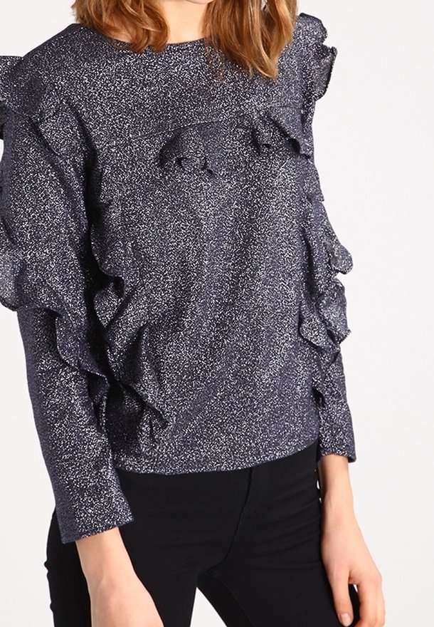Femme portant un haut à manches longues bleu marine pailleté avec des détails à volants sur le devant et les manches, associé à un pantalon noir.