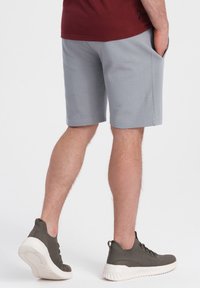 Pantalones cortos grises de tela suave, con bolsillos laterales y un corte relajado. Llevados con zapatillas deportivas de color verde oscuro con suela blanca.