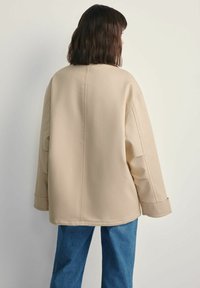 Osoba o ciemnych włosach sięgających do ramion, ubrana w oversize'ową beżową kurtkę i niebieskie dżinsy, stoi plecami do prostej jasnej tła.