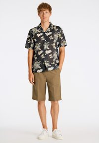 Camicia nera a maniche corte con fantasie floreali verdi e bianche, abbinata a pantaloni shorts kaki e sneakers bianche. Il modello si trova di fronte a uno sfondo neutro.