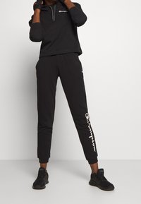 Sudadera negra con capucha y logo blanco en el pecho, combinada con joggers negros ajustados con logo blanco en la pierna, y zapatos deportivos negros.
