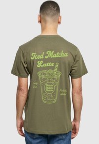 Oliven grøn t-shirt med et stort, lys grønt grafisk motiv af en iset matcha latte med tekst, rund halsudskæring og korte ærmer.