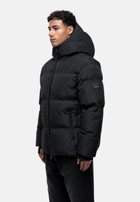 Black puffer jacket with a high collar and oversized hood, featuring zippered side pockets, quilted texture, and a logo patch on the sleeve.