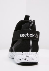 Baskets de sport noires avec tige en mesh et en suède, semelle blanche mouchetée, logo Reebok sur la sangle et languette à rayures noir et blanc.