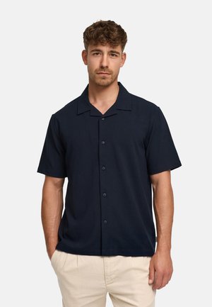 Homme aux cheveux bouclés portant une chemise noire à manches courtes boutonnée et un pantalon beige, debout devant un fond clair uni.