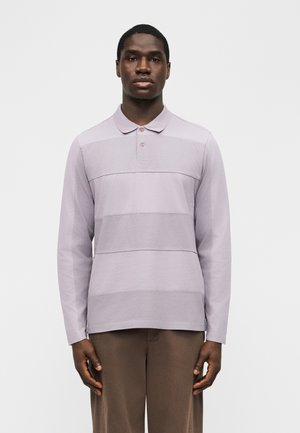 Homme debout portant un polo à manches longues violet clair et un pantalon marron devant un fond uni.