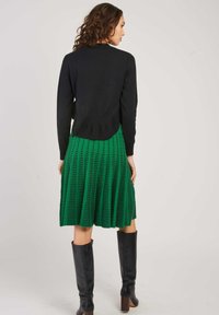Pull en maille noire avec un ourlet côtelé, associé à une jupe plissée verte avec un motif en grille texturé, et des bottes noires à hauteur de genou.