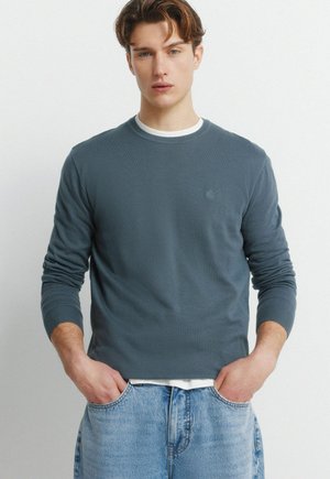 Młody mężczyzna o krótkich brązowych włosach, ubrany w ciemnoniebieski sweter z długim rękawem na białą koszulę oraz jasnoniebieskie dżinsy, z rękami w kieszeniach, na neutralnym tle.