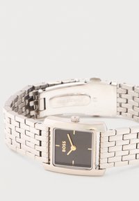 Ατσάλινο ασημί ρολόι χειρός με ορθογώνιο μαύρο καντράν, χρυσά δείκτες και λογότυπο BOSS στο πρόσωπο, εμφανιζόμενο με το κούμπωμα μερικώς ανοιχτό.